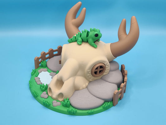 Decorative figurine of a deer skull with a lizard on top, surrounded by greenery and rocks